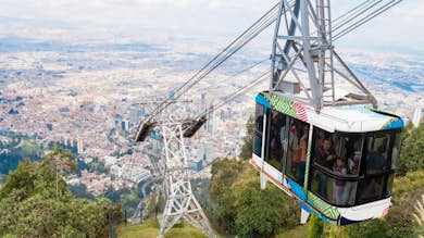 Colorido teleférico que asciende sobre un denso paisaje urbano, con los pasajeros dentro y una exuberante vegetación debajo.. Abrir en galería a pantalla completa.