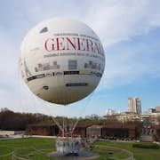 Un gran globo aerostático amarrado con texto publicitario y diseños de paisajes urbanos, posado en tierra en un parque con personas y edificios al fondo.