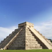 Una antigua pirámide de piedra con escalones empinados bajo un cielo azul claro, rodeada de vegetación.