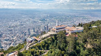 Vista aérea de un paisaje urbano con una gran iglesia en lo alto de una colina rodeada de exuberante vegetación y numerosos edificios en la parte inferior.. Abrir en galería a pantalla completa.